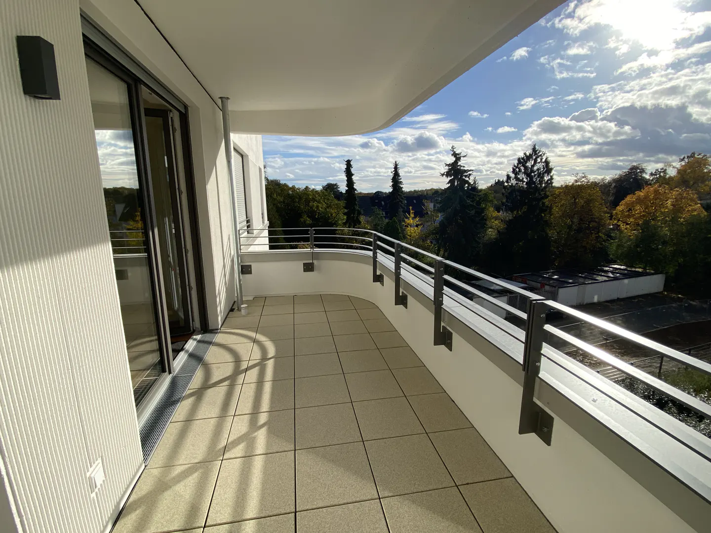 A modern balcony with beige tiles, a metal railing, and a view of trees under a blue sky with clouds.