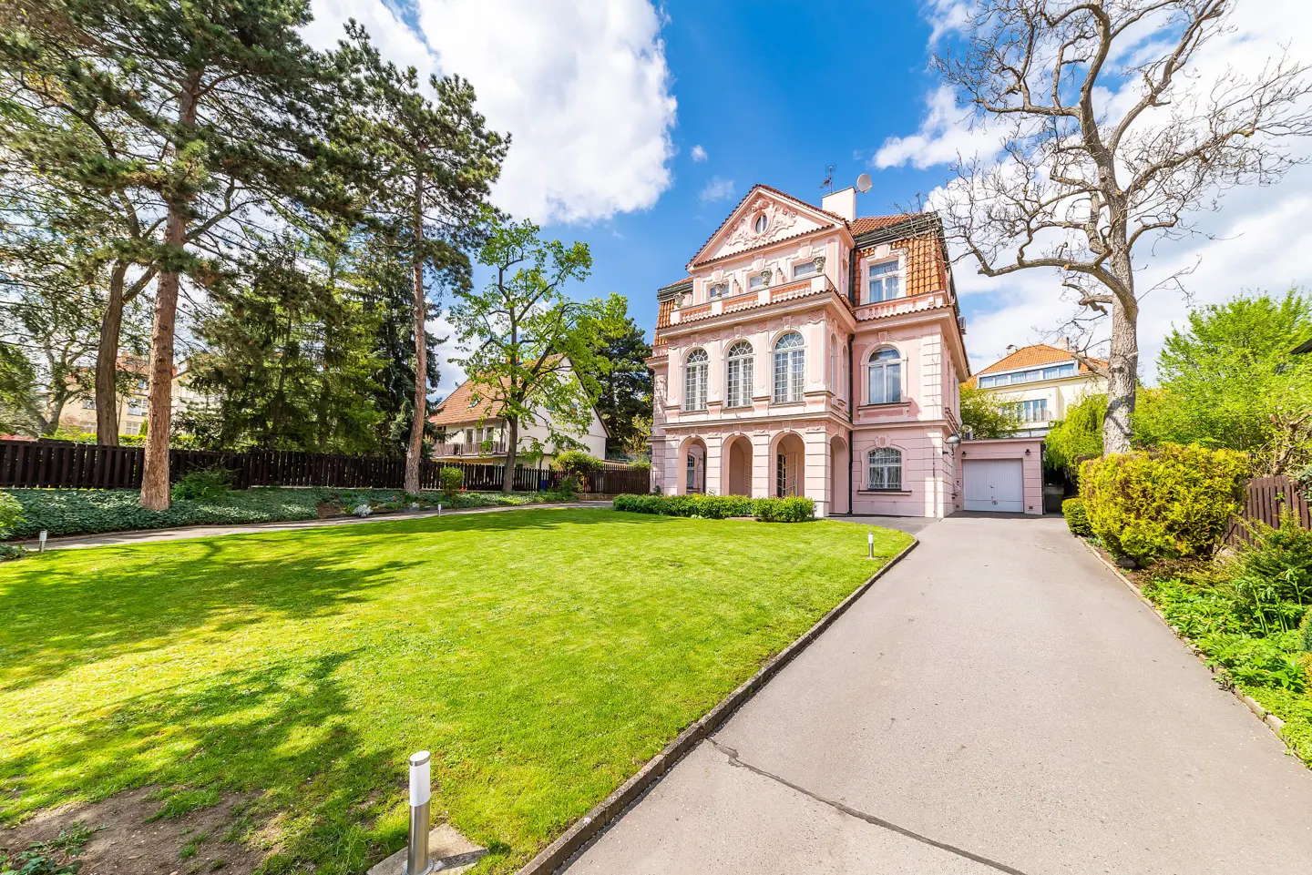 Exterior view of a two-story pink house with a green lawn, driveway, and trees under a blue sky with white clouds.