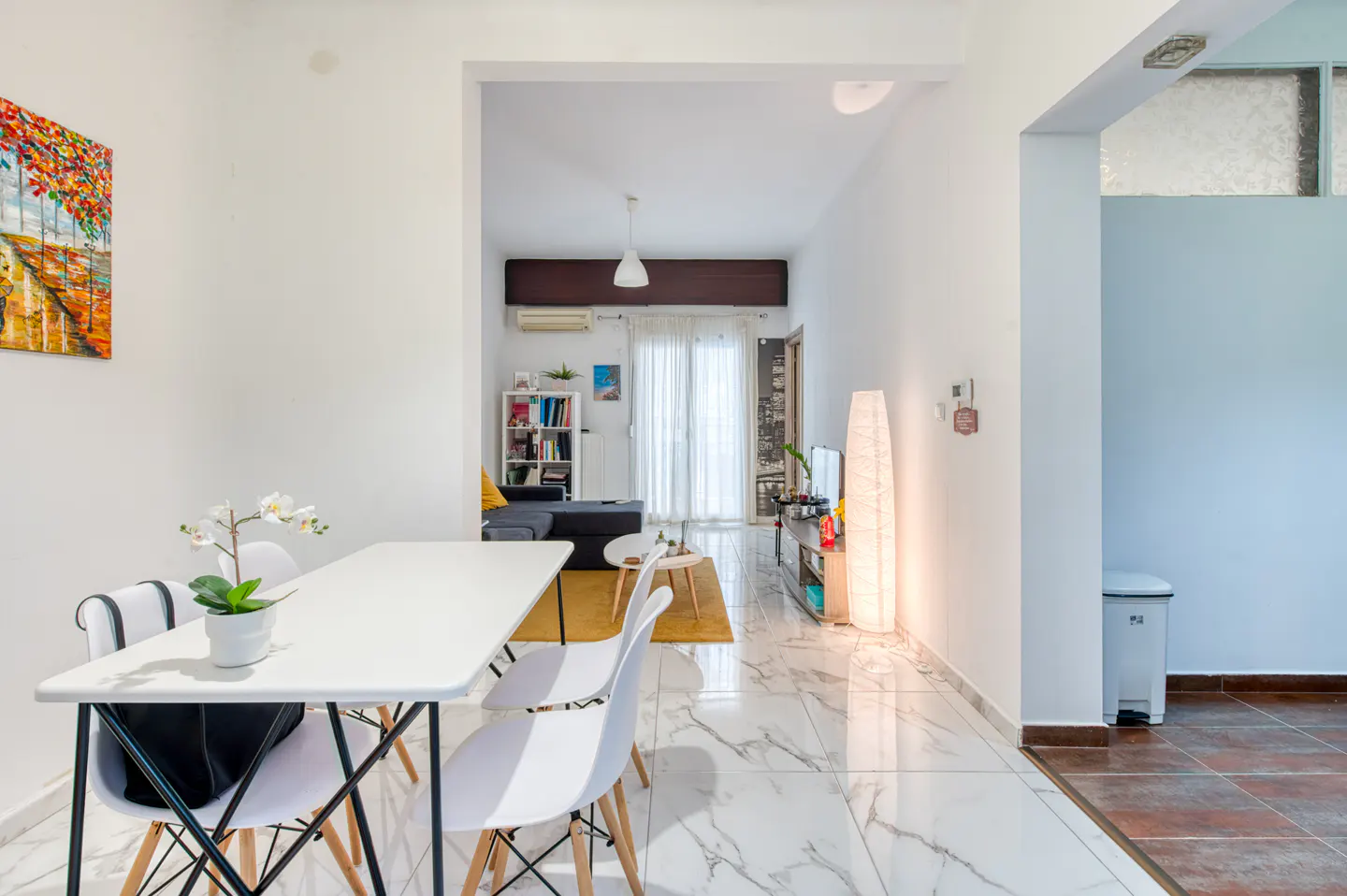 Bright, open-concept apartment interior. A white table with chairs sits near a colorful painting. Living room visible in the background.