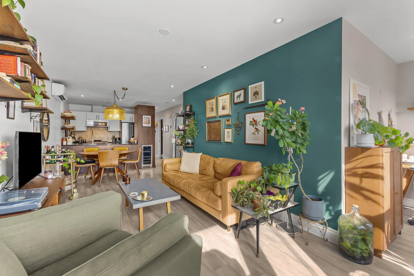 A living room with a green accent wall, a mustard-colored sofa, and various plants. The kitchen and dining area are visible in the background.