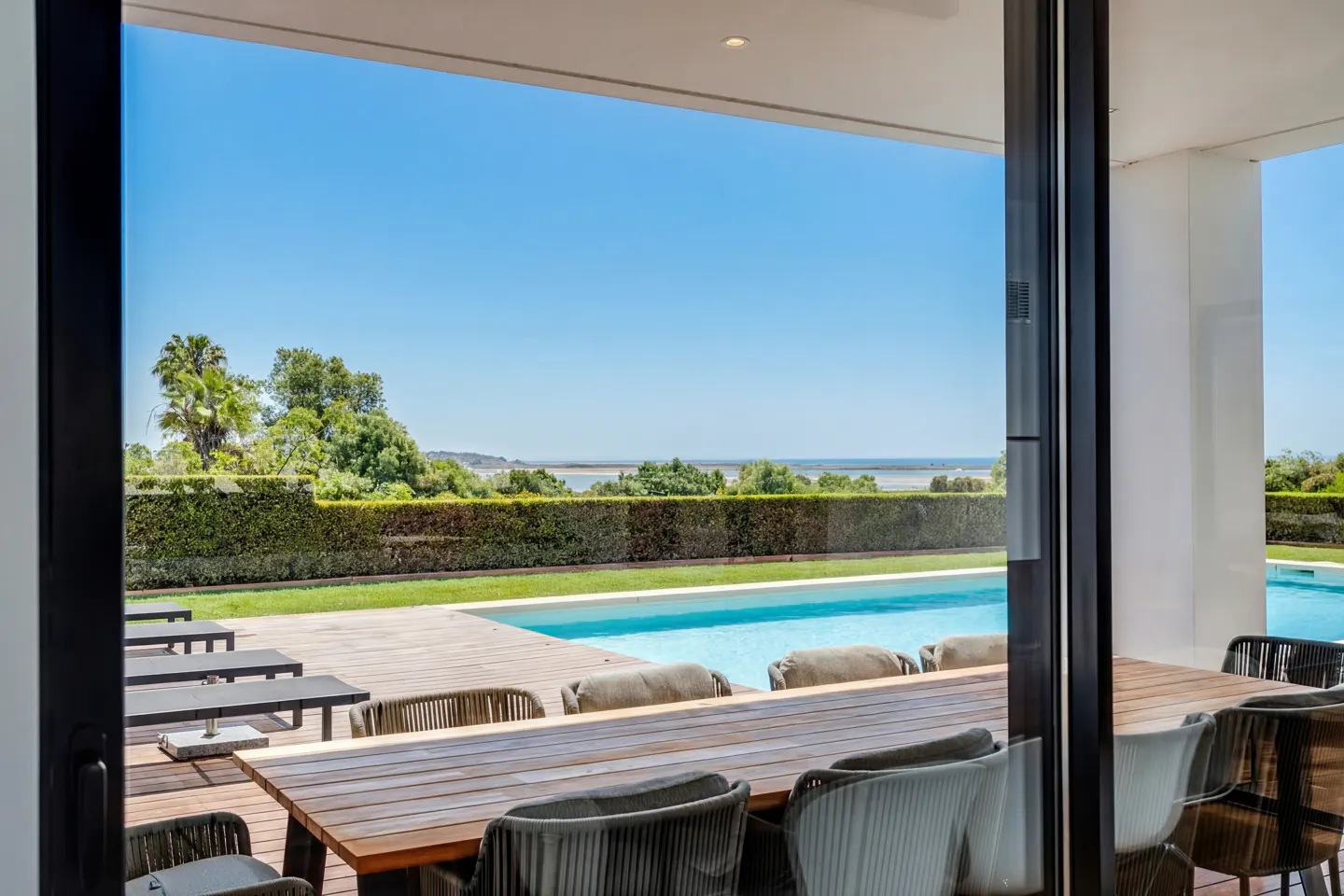 View from a patio with a wooden table and chairs, overlooking a pool, green hedge, and blue sky.