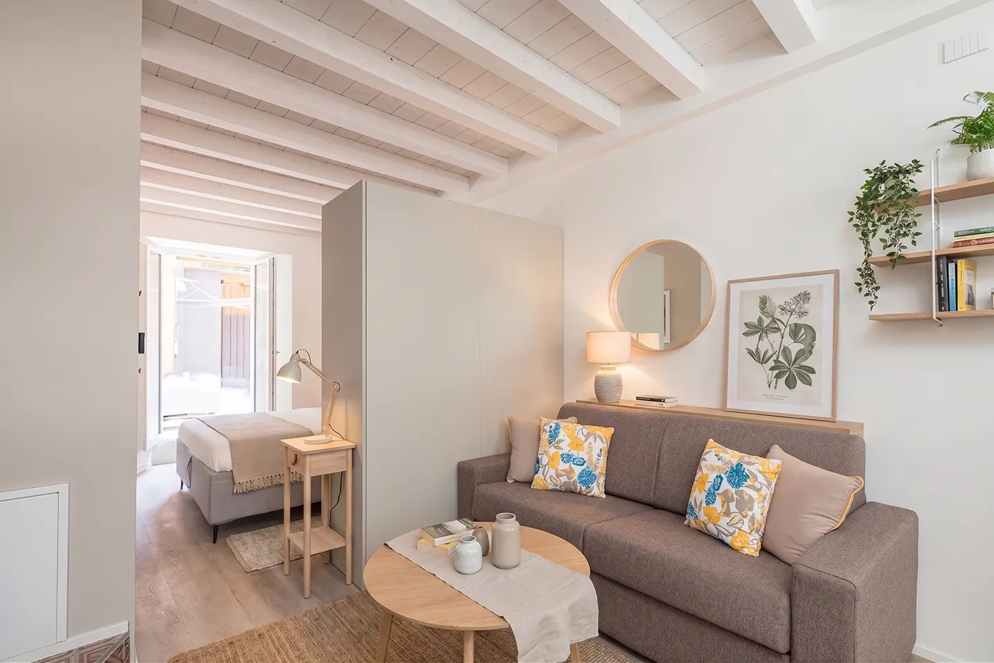 Modern studio apartment with a brown sofa, round coffee table, and a bedroom area visible behind a partition wall.
