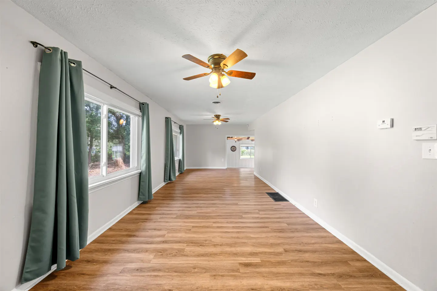 Long, empty room with light wood-look flooring, white walls, two ceiling fans, and green curtains over large windows.