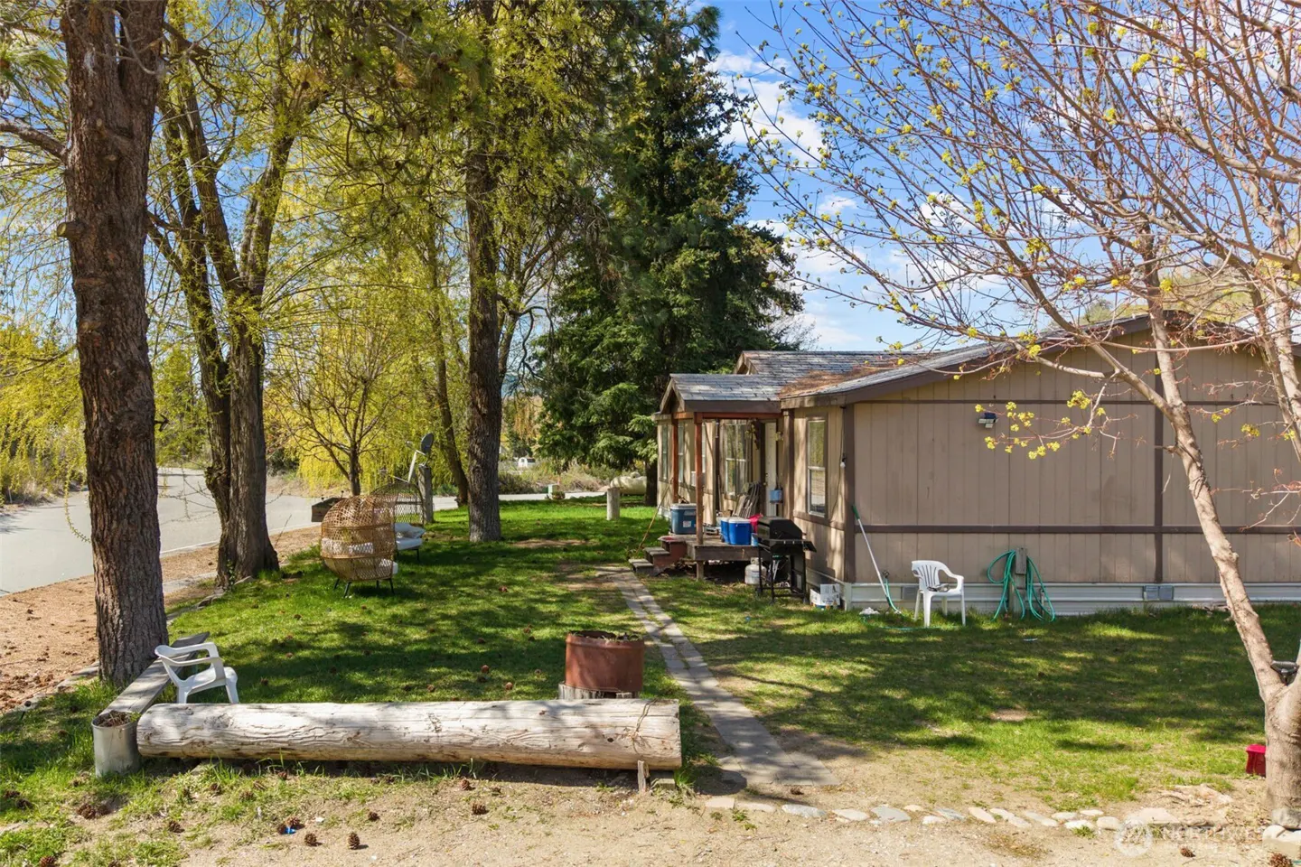 A tan mobile home with a porch and green lawn, trees, and a blue sky.