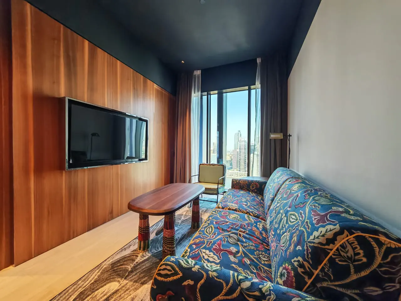 A modern living room with a blue floral sofa, wooden table, and a flat-screen TV mounted on a wooden wall. A large window offers a city view.