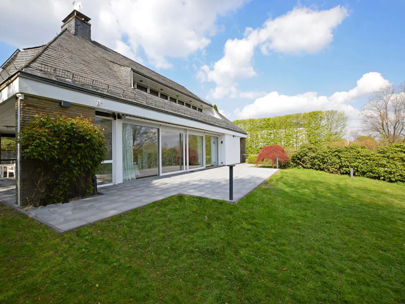 Modern house exterior with gray roof, large sliding glass doors, gray patio, and green lawn under a blue sky with white clouds.
