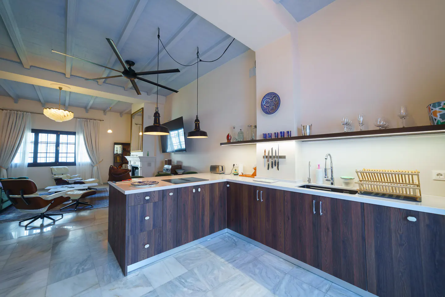 Open-concept kitchen and living room with marble floors, dark wood cabinets, and modern furniture. A large ceiling fan hangs above.