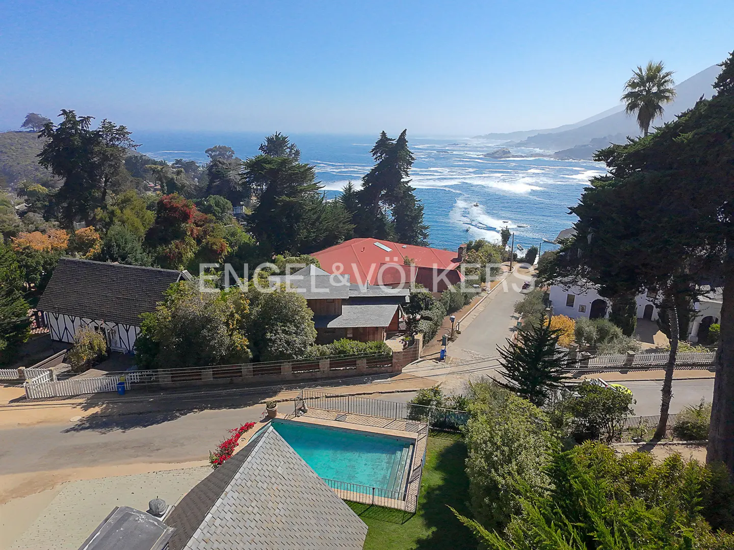 Real estate view of homes with pools, trees, and ocean views under a blue sky. Engel & Völkers logo is visible.