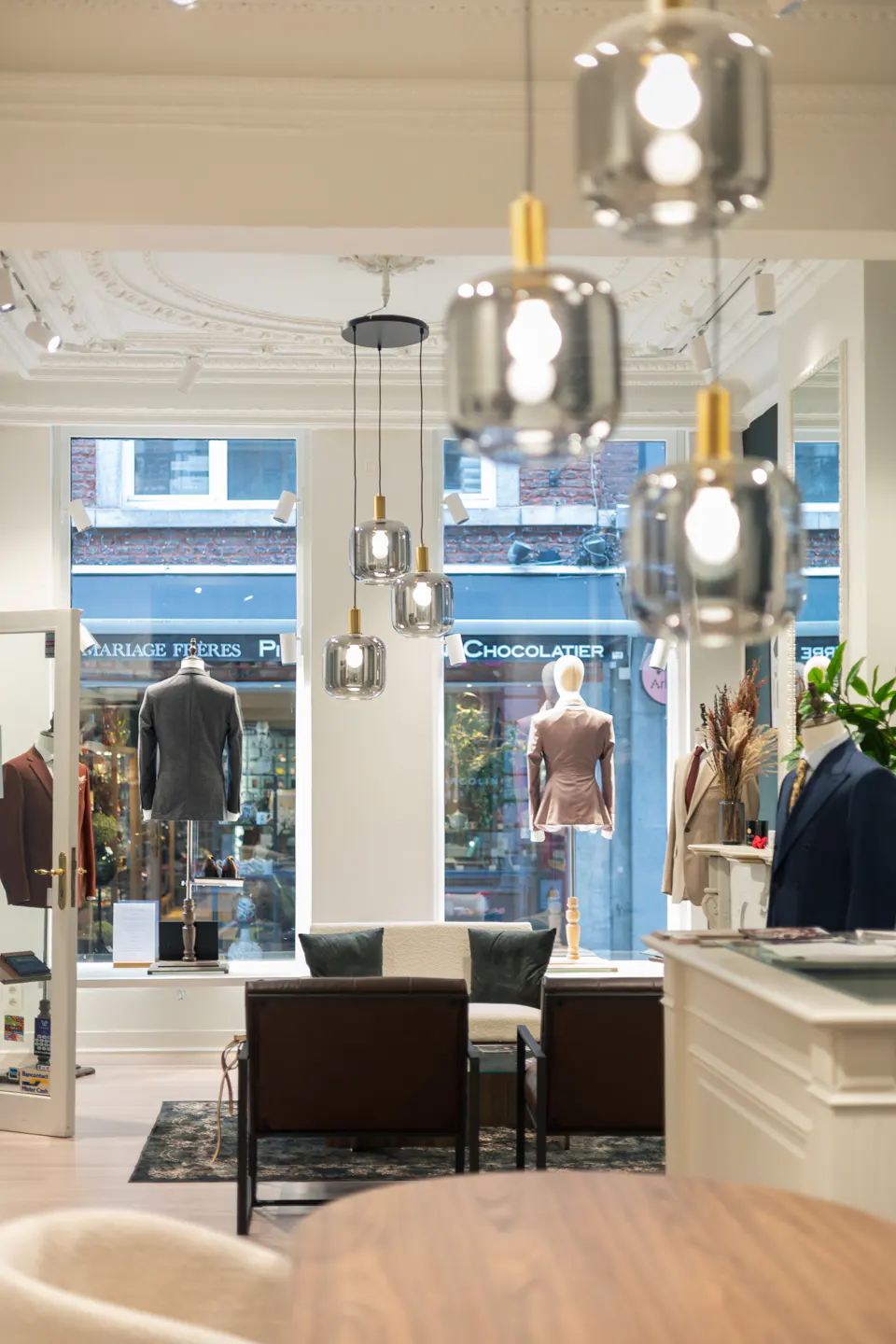 Upscale clothing store interior with suits on mannequins, modern lighting, and a seating area. The color palette is neutral with pops of color from the clothing.