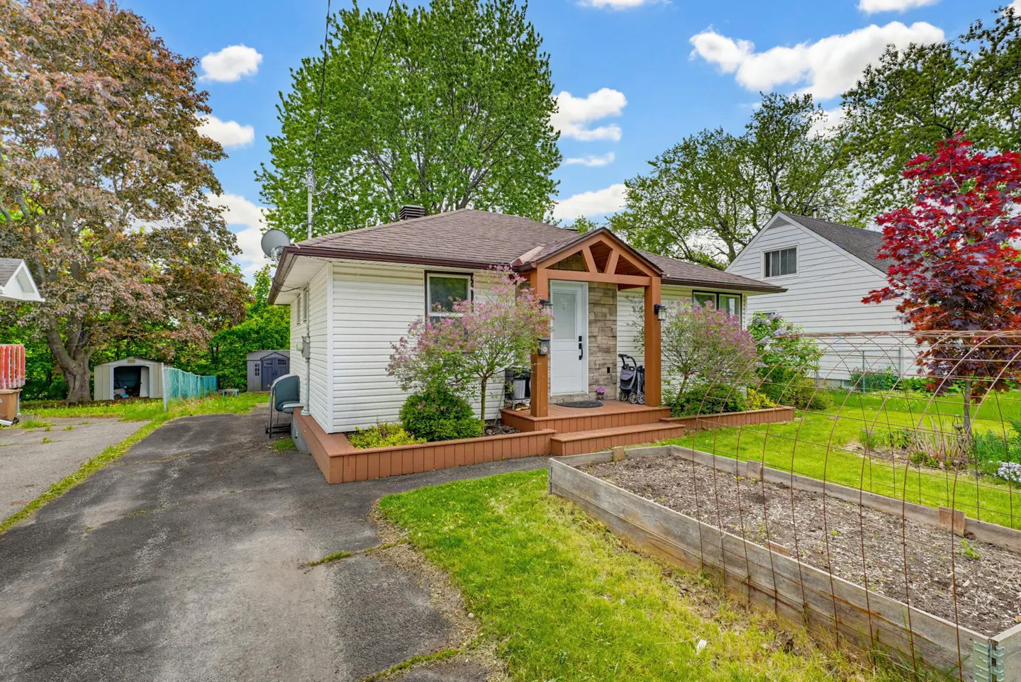 White bungalow with a wooden porch, stone accents, a paved driveway, and a raised garden bed in the front yard.