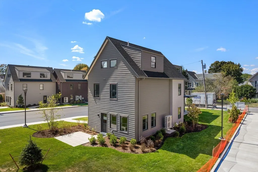 Two-story gray house with a black roof, green lawn, and blue sky. Other houses are visible in the background.