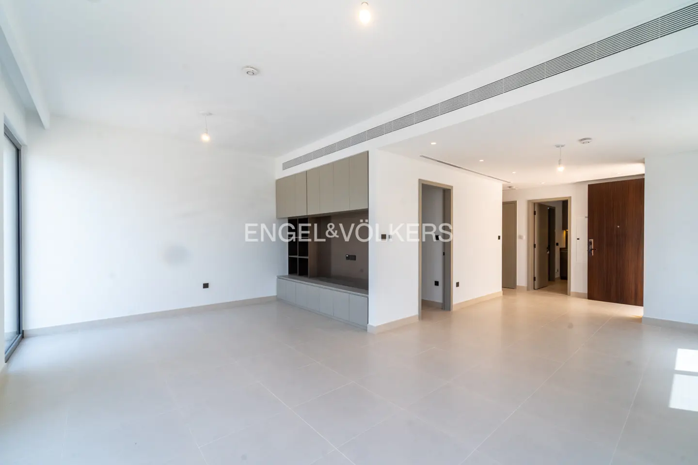 Bright, empty living room with white walls, beige tile floor, and built-in gray cabinets. Doors lead to other rooms. Engel & Völkers logo visible.