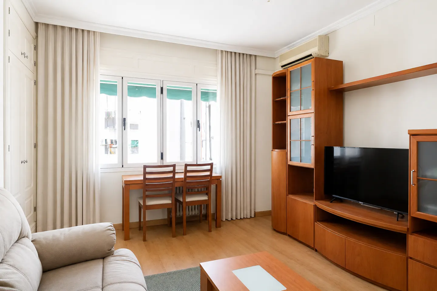 Living room with a beige sofa, wooden table and chairs by a window with white curtains, and a wooden entertainment center with a TV.