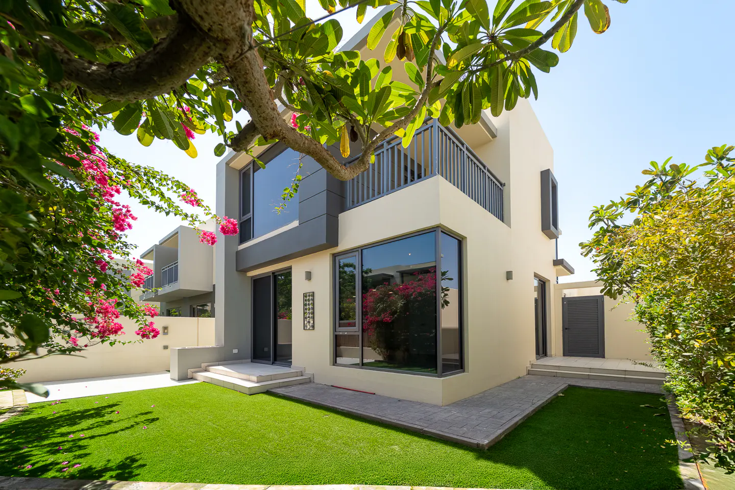 Two-story modern house with a balcony, large windows, and a green lawn, framed by trees and pink flowers.