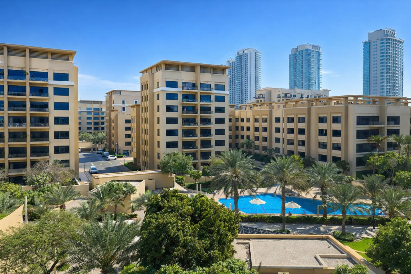 Apartment complex with a pool. Tan buildings with dark windows, palm trees, and blue pool with umbrellas. Tall buildings in the background.