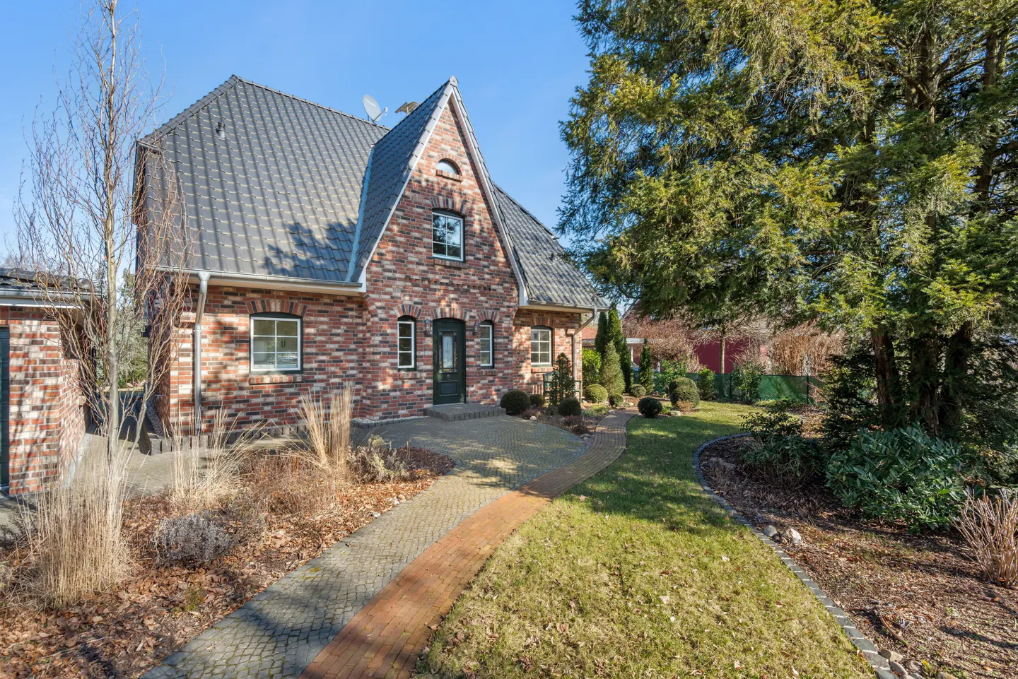 A two-story brick house with a gray roof and a brick walkway leading to the front door. A large tree is on the right side of the house.