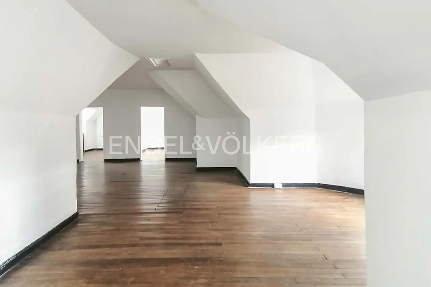 Attic room with white walls, a brown wood floor, and a skylight. Doorways lead to other rooms. Engel & Völkers logo visible.