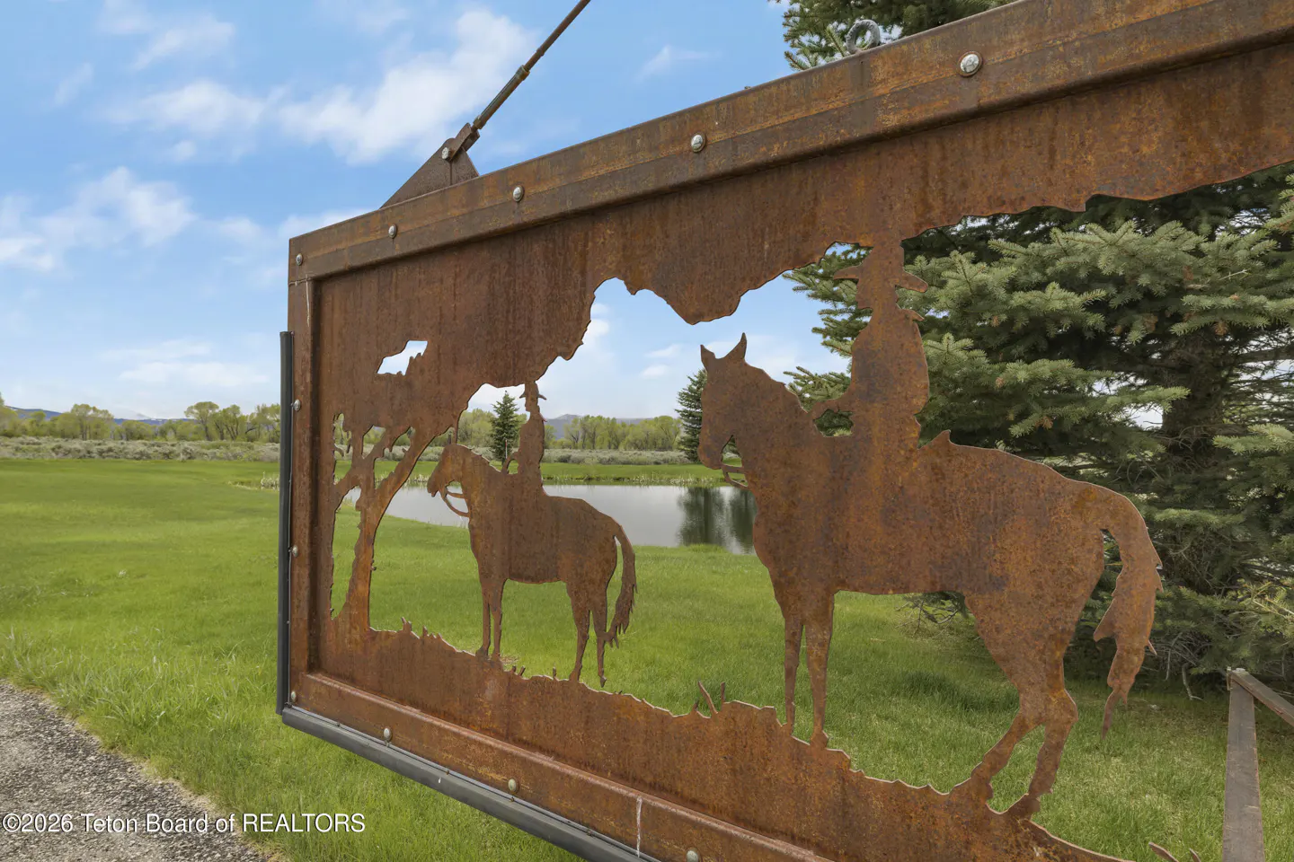 Rusty metal gate with cut-out silhouettes of cowboys on horseback, overlooking a green field and pond.