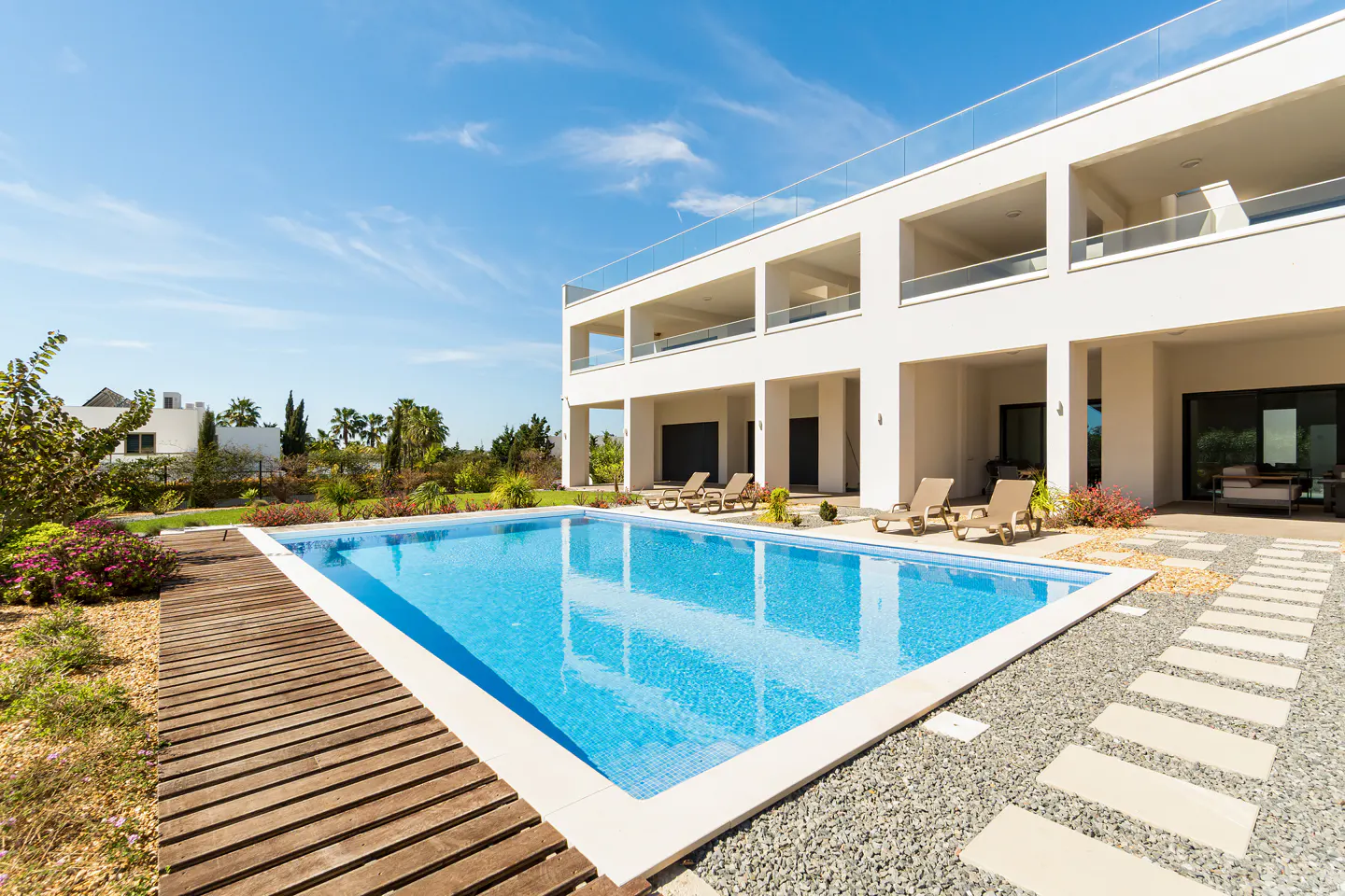 A modern white house with a blue tiled pool, wooden deck, and lounge chairs under a sunny sky.