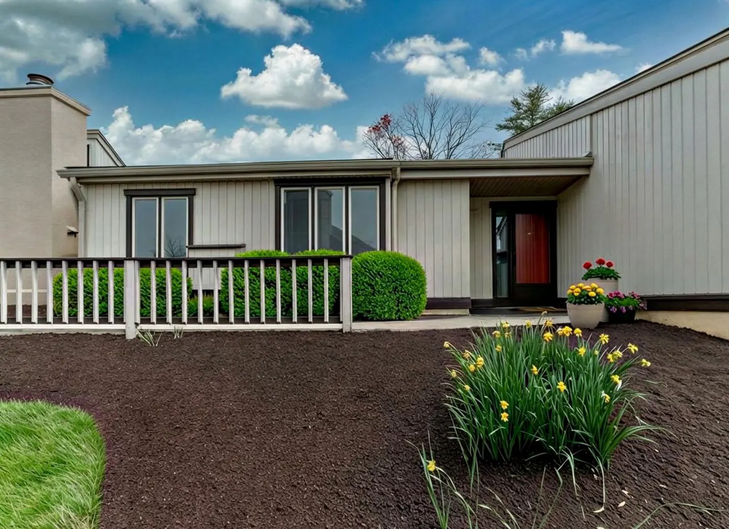 A one-story house with a red door, white fence, green bushes, and yellow flowers.