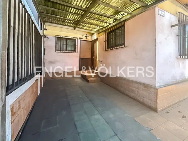 Covered patio with a metal roof, pink walls, barred windows, and an open door with steps. The Engel & Völkers logo is superimposed.