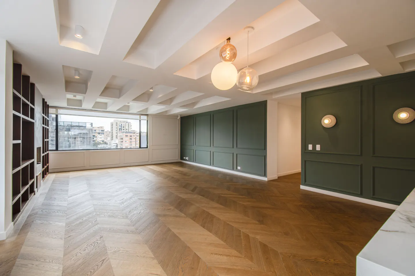 Bright, empty room with herringbone wood floors, green paneled walls, and a large window with a city view. Modern lighting fixtures hang from the decorative ceiling.