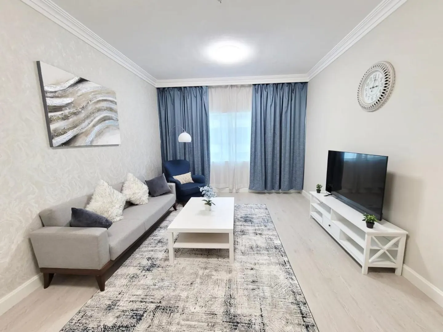 A bright living room with a gray sofa, white coffee table, blue curtains, and a TV on a white stand.