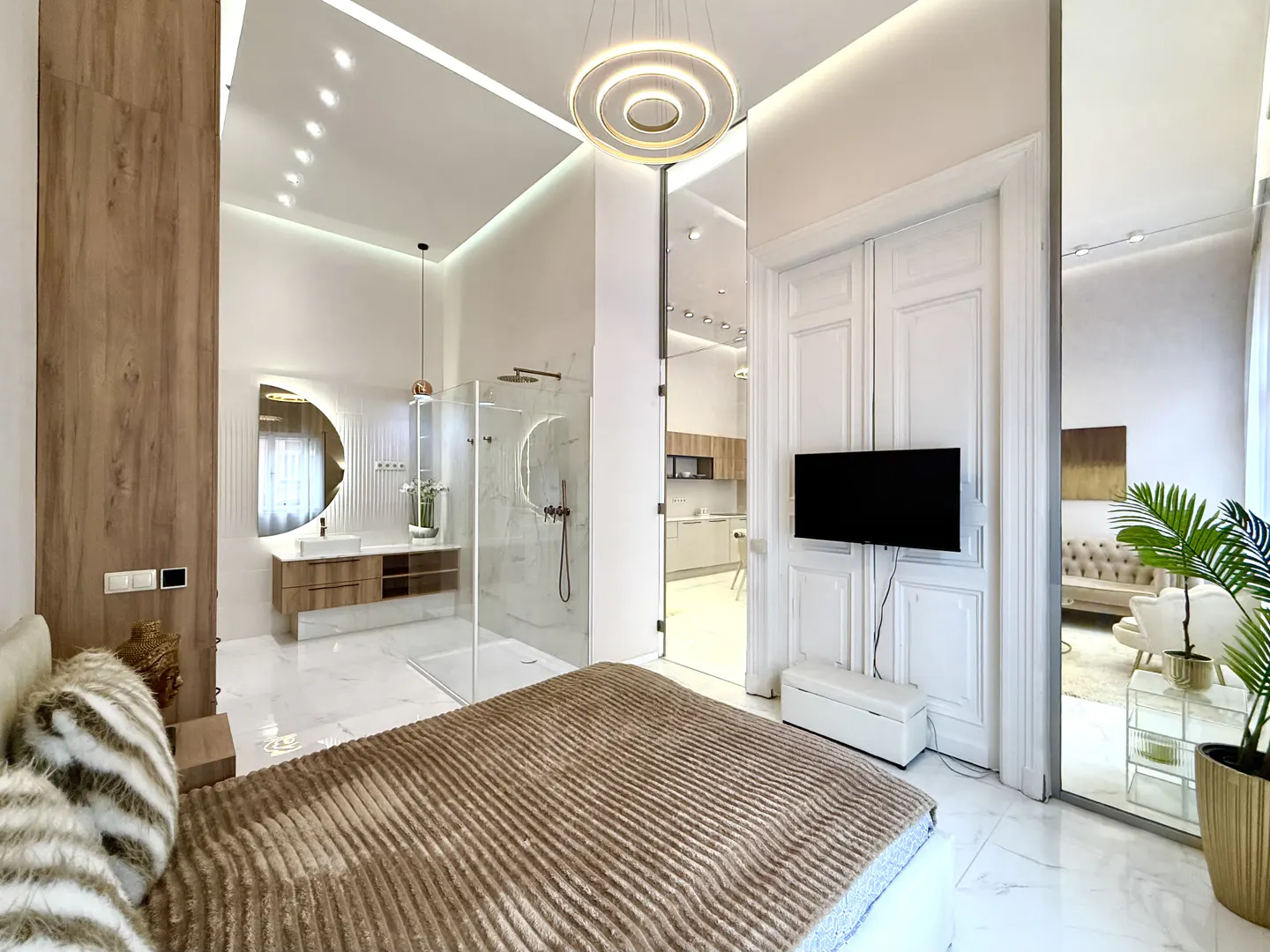 A modern bedroom with a brown striped bedspread, white marble floors, and an open bathroom with a glass shower. A TV is mounted on a white door, and a kitchen is visible in the background.