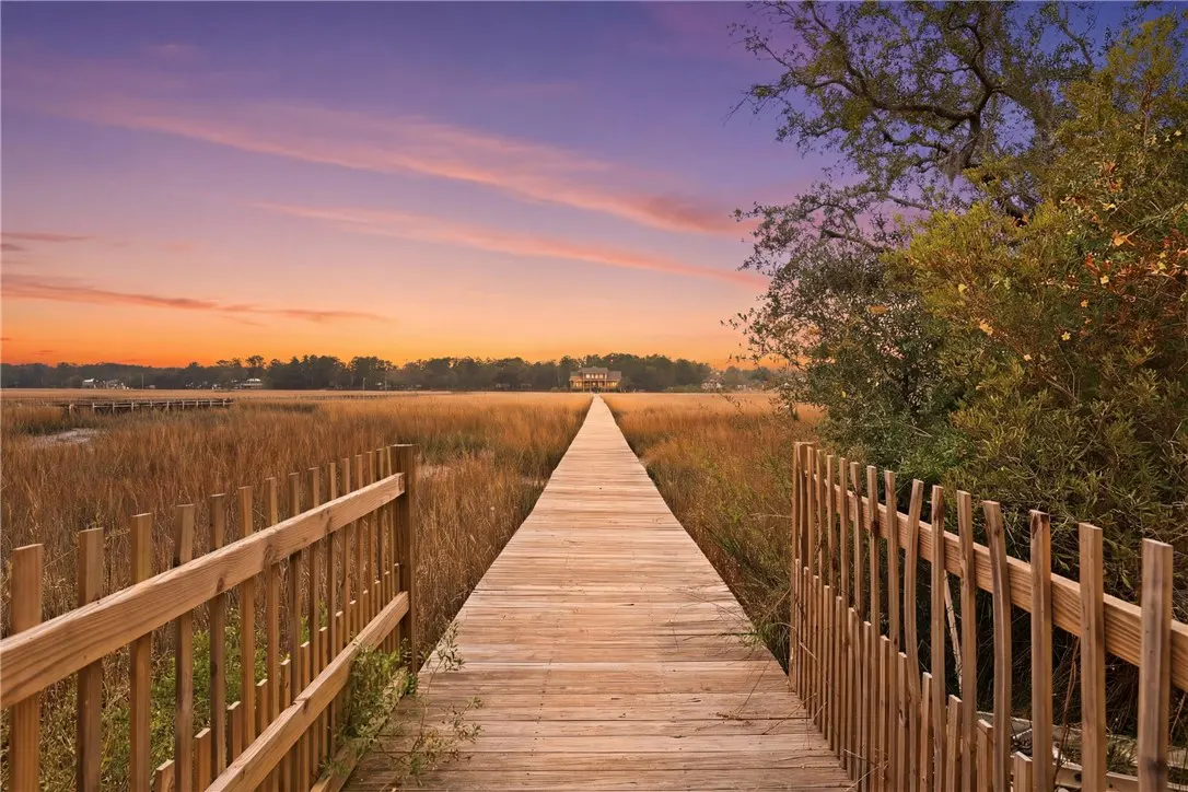 Wooden boardwalk through marsh grass leads to a house under a colorful sunset sky. Fences line the boardwalk.