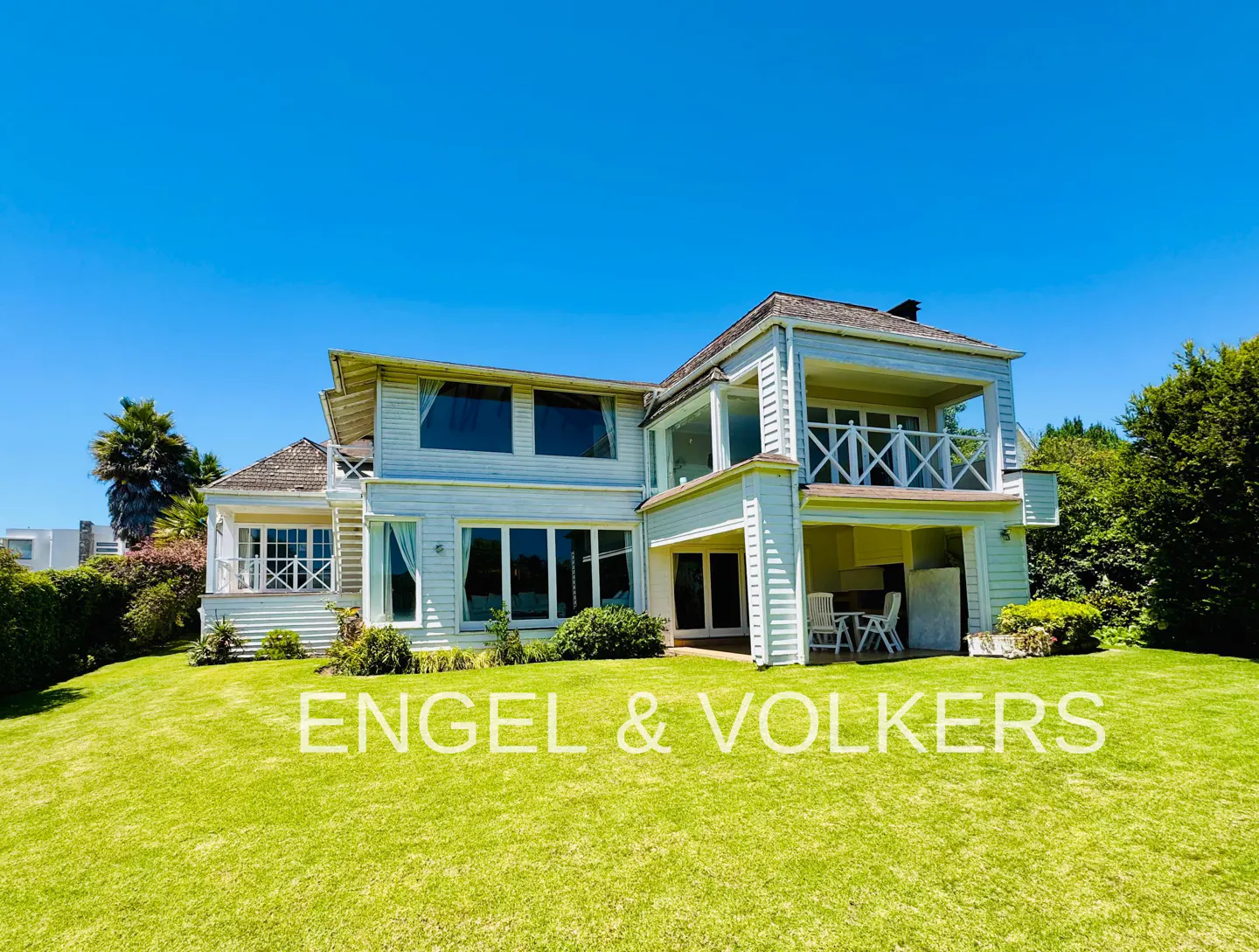 Two-story white house with a balcony and large windows on a green lawn under a clear blue sky. Engel & Volkers logo is superimposed.