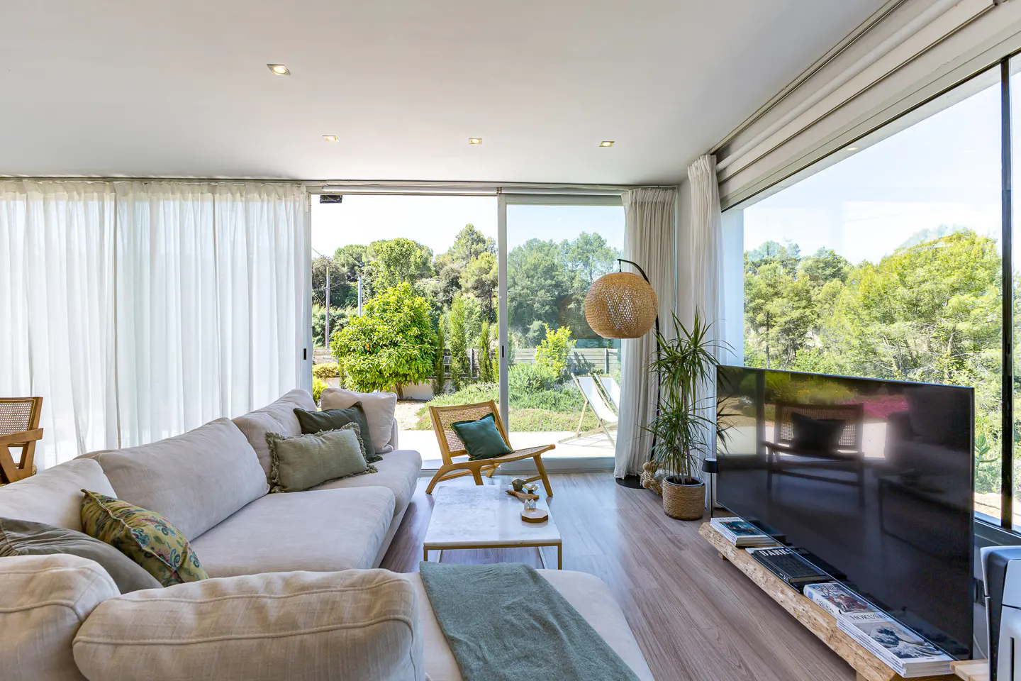 Bright living room with a large beige sofa, wood floors, and floor-to-ceiling windows overlooking a green garden. A rattan chair and a TV stand add natural elements.