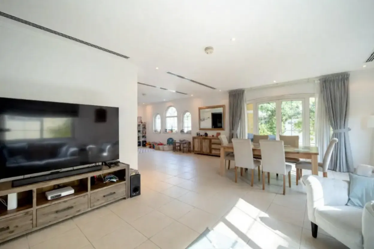 Bright, open-plan living and dining area with white walls and tile floors. A large TV sits on a wooden console. Dining table with white chairs near a window.