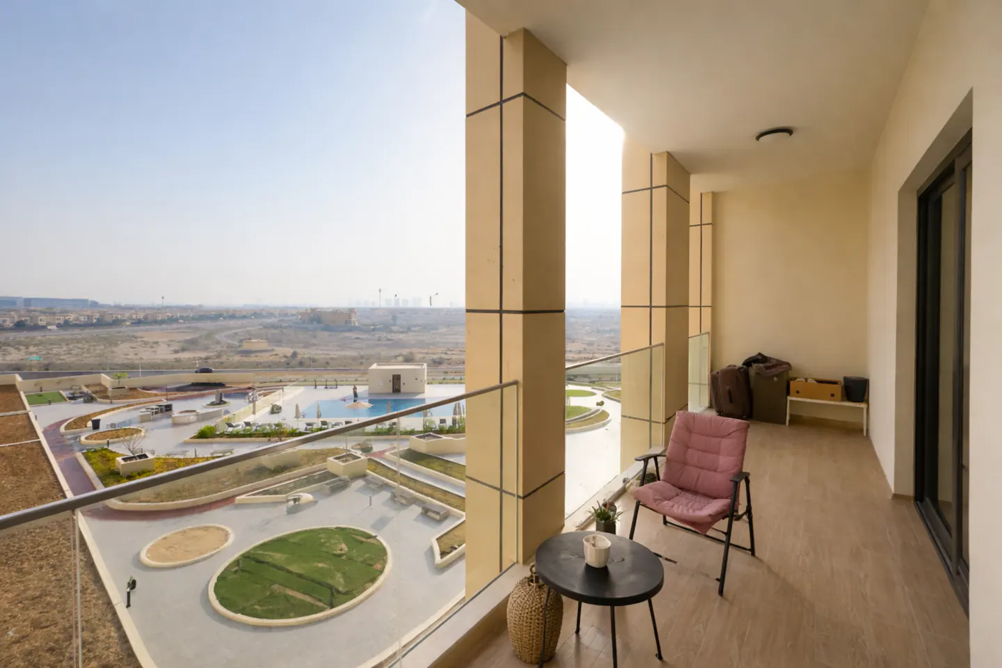 Balcony view of a pool and desert landscape. A pink chair and black table sit on the balcony. Luggage is stacked near the sliding glass door.
