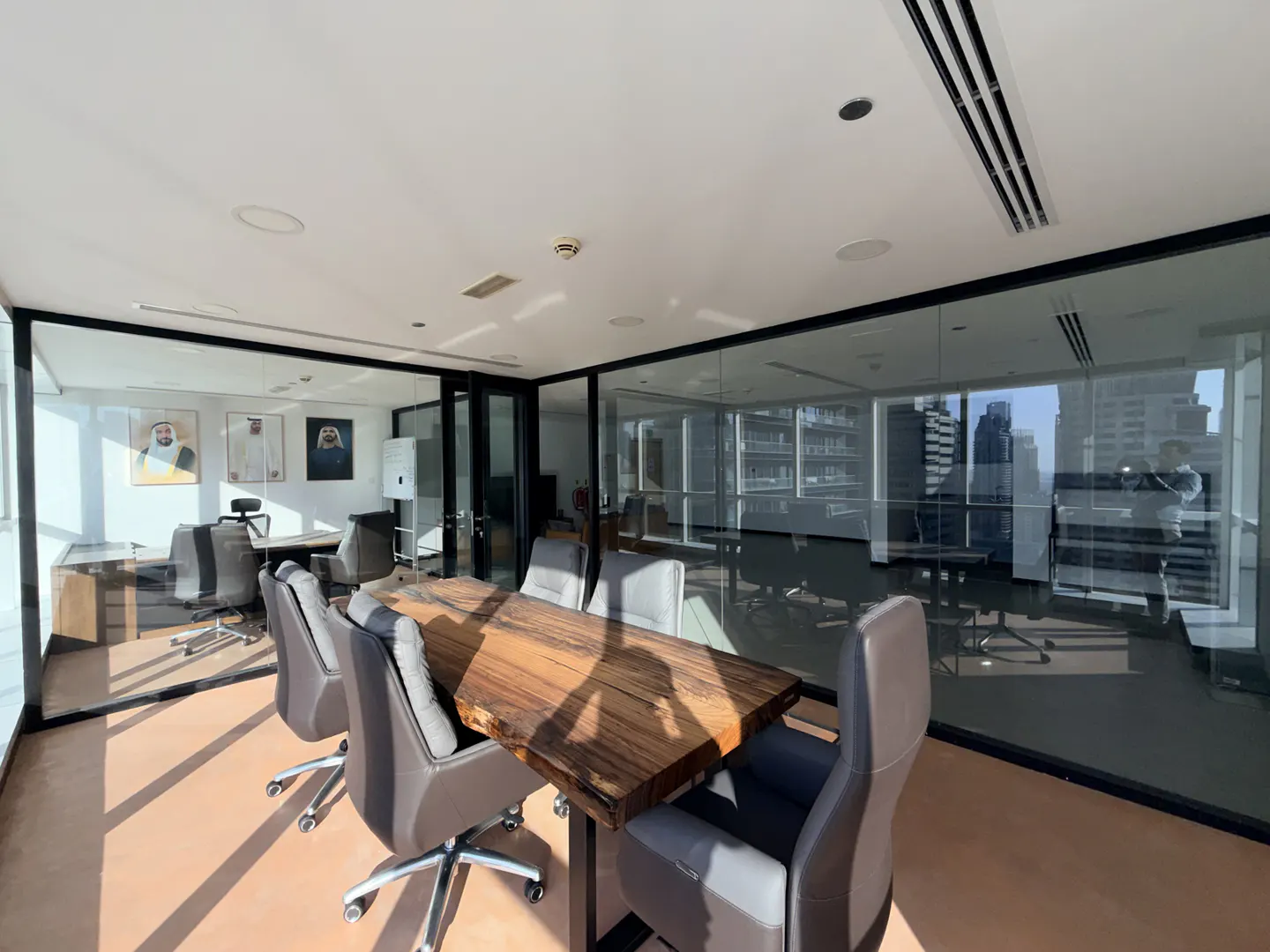 A modern office space with a large wooden conference table surrounded by gray chairs, glass walls, and city views.