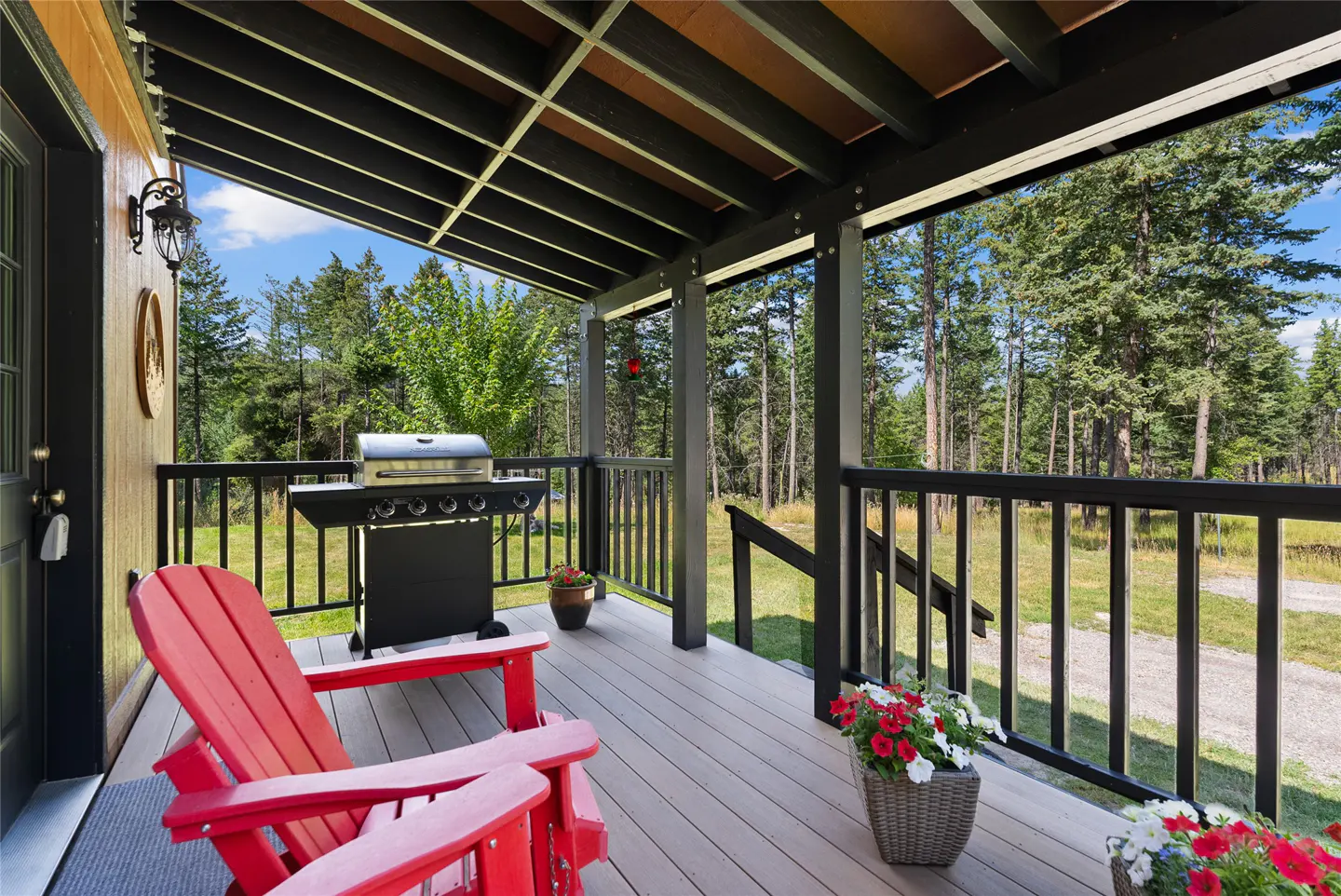 Covered porch with red Adirondack chairs, grill, and flower pots. Black railings overlook a forest.