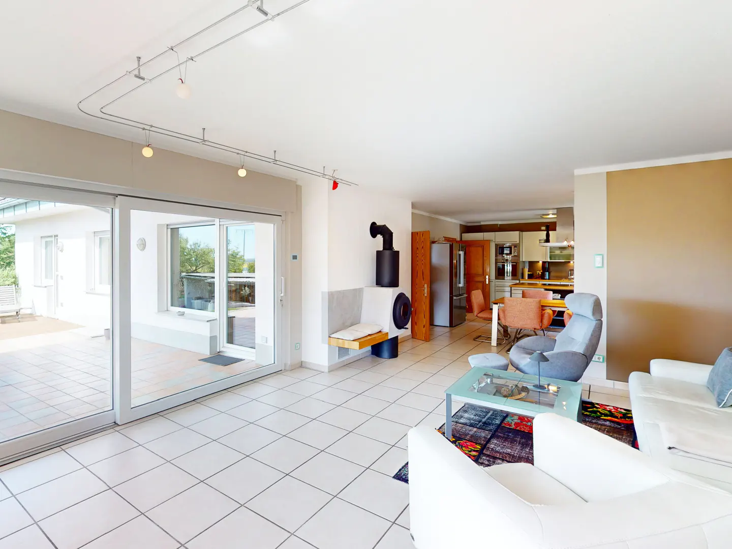 Bright, open living room with white tile floor, white sofas, and a modern fireplace. Sliding glass doors lead to a patio. Kitchen in background.