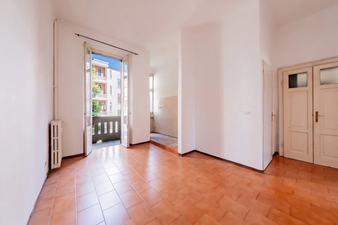 An empty room with terracotta tile floors, white walls, and an open balcony door. A radiator is on the left wall. Double doors are on the right.