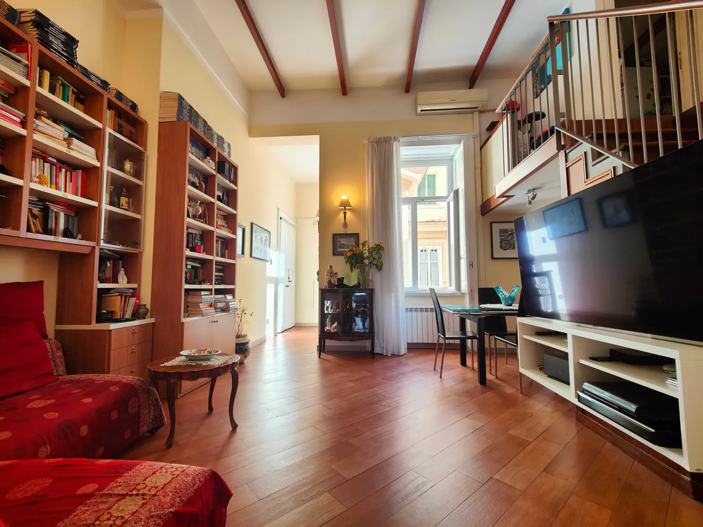 Living room with wood floors, red sofa, and bookshelves. A dining table sits near a window, and a loft is visible above.