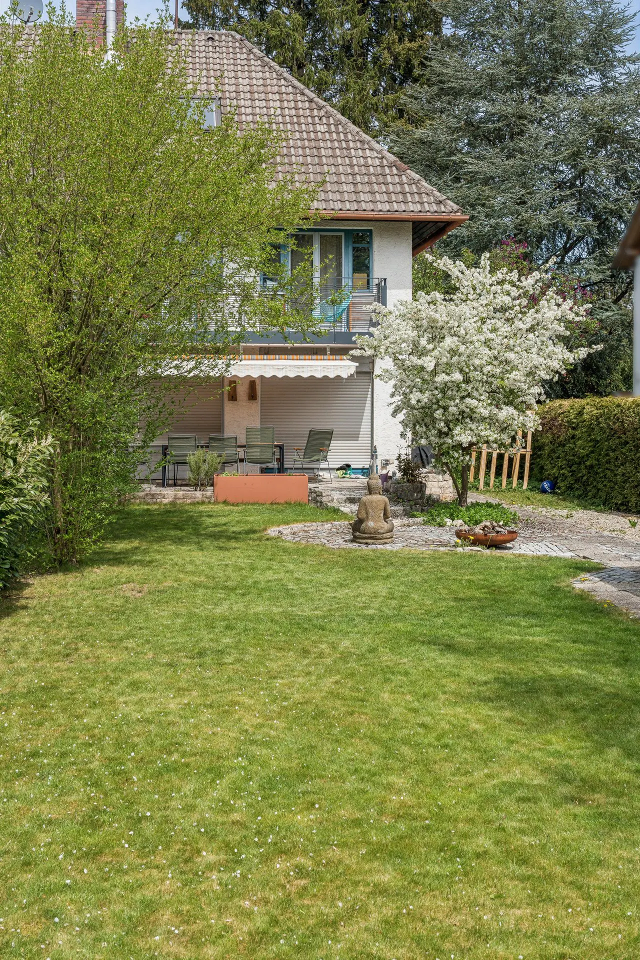 A house with a green lawn, trees, and a patio with outdoor furniture. A Buddha statue sits near a flowering tree.