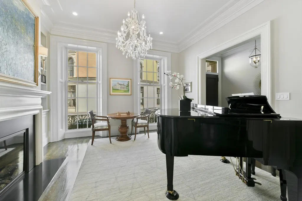Elegant living room with a black grand piano, fireplace, and a round table with two chairs near windows. A crystal chandelier hangs from the ceiling.
