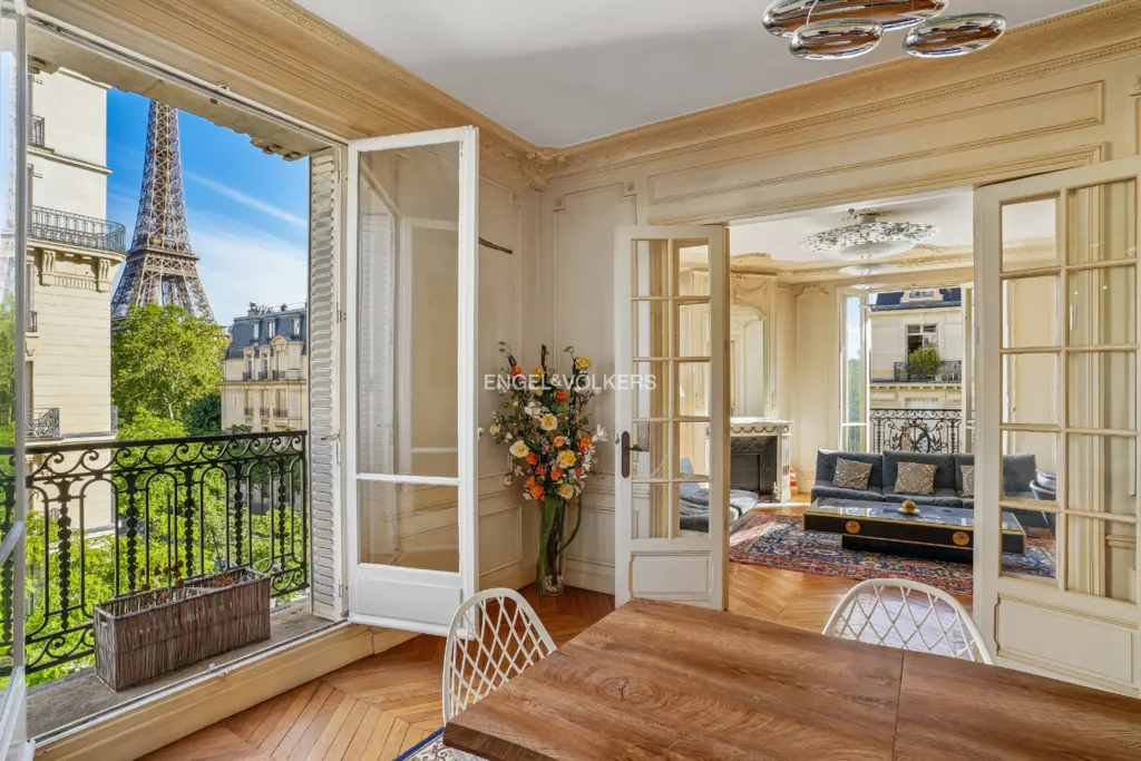 Interior view of a Paris apartment with a balcony overlooking the Eiffel Tower. A wooden table and chairs are in the foreground.