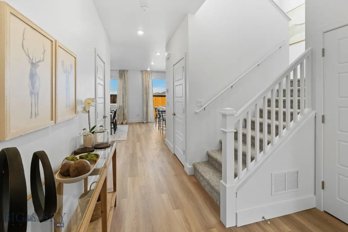 Bright, modern entryway with light wood floors, white walls, and a staircase. Deer artwork hangs above a console table with decorative objects.