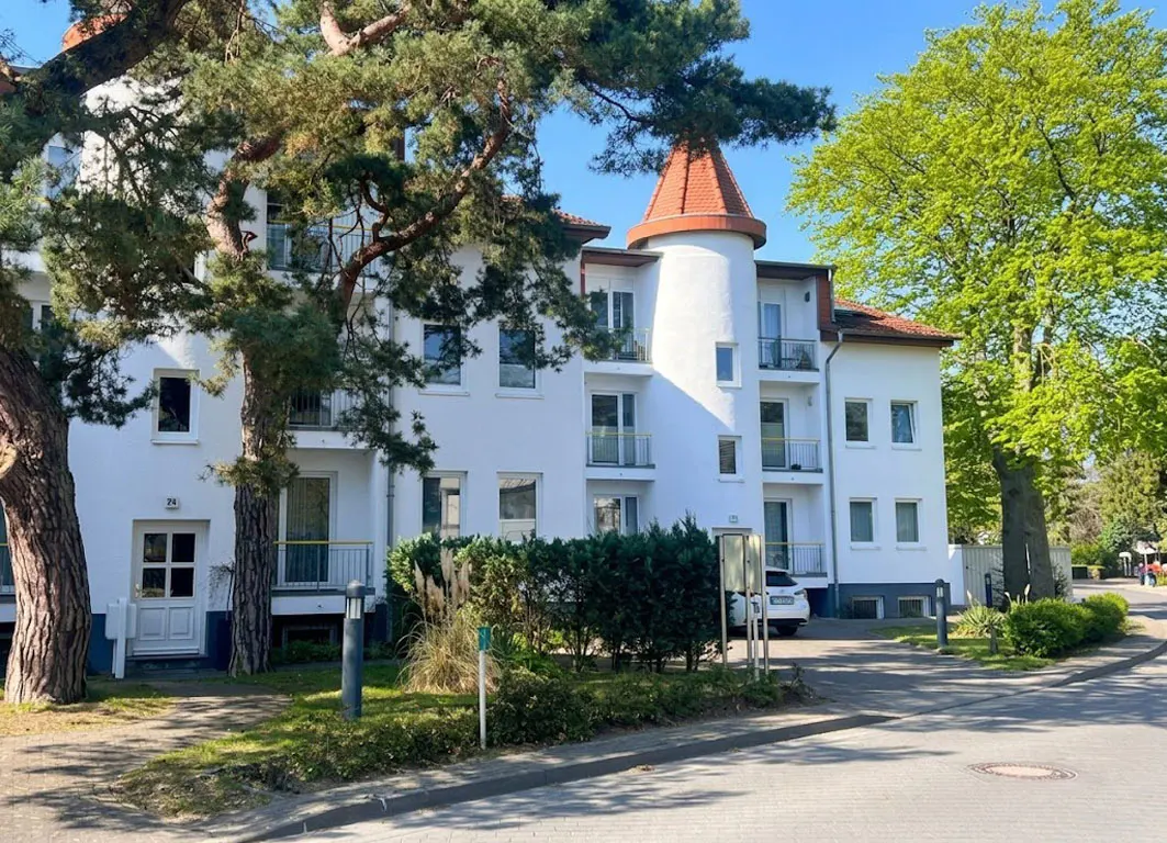 Exterior of a three-story white apartment building with balconies and a turret with a red roof. Trees surround the building.