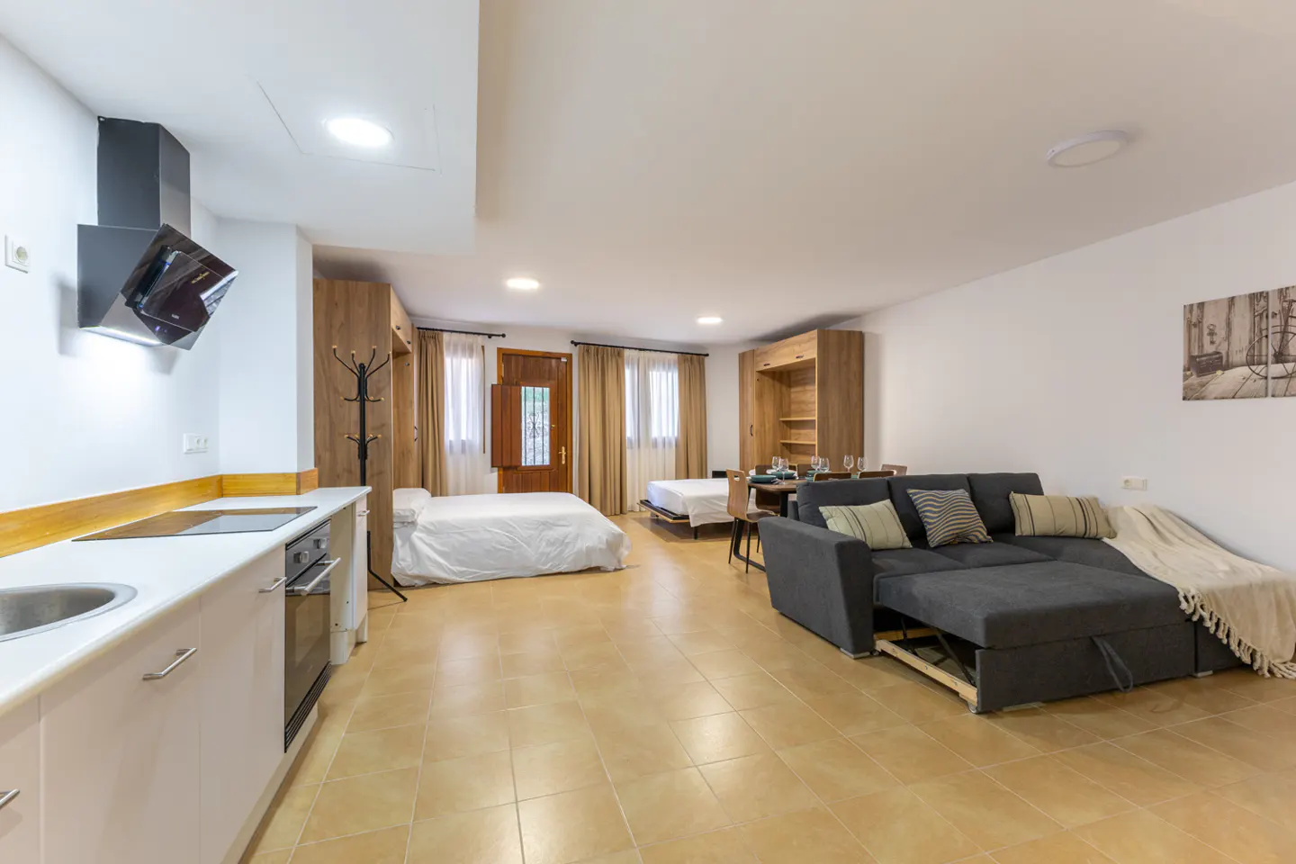 Bright studio apartment with a kitchen, two beds, a dining table, and a gray sofa with pillows. The walls are white and the floor is tiled.