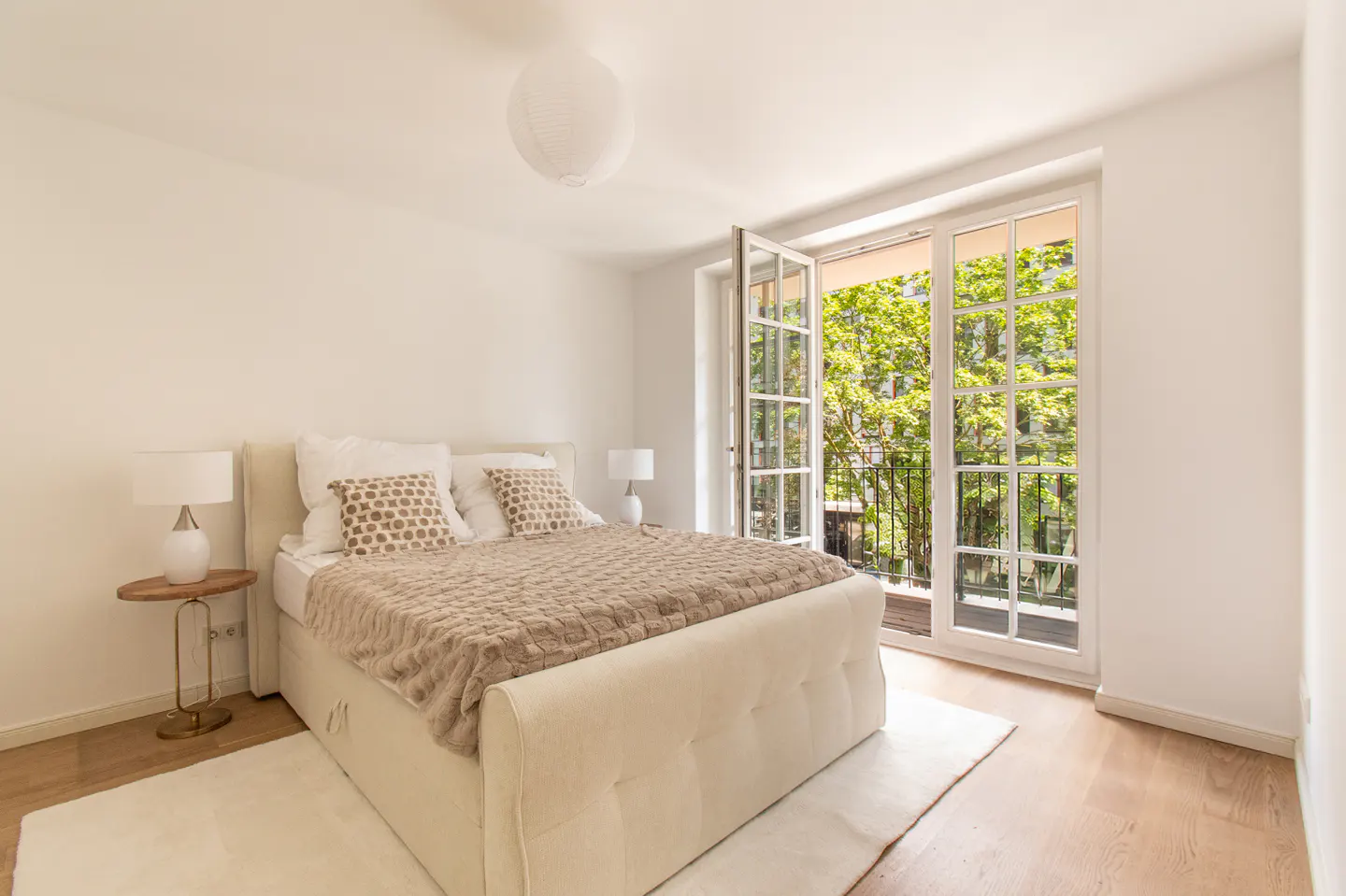 Bright bedroom with a tufted cream bed, beige throw, and open French doors revealing lush green trees outside.
