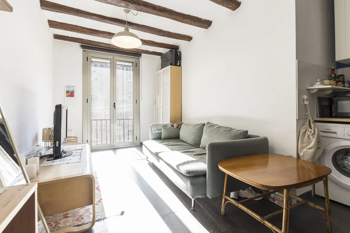 Bright living room with white walls, wood beams, and a gray sofa. A table sits in front of the sofa, and a washing machine is visible in the background.