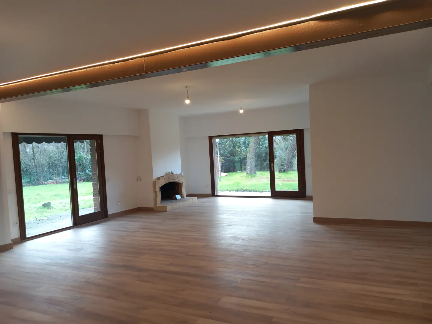 Bright, empty living room with wood floors, white walls, and a stone fireplace. Sliding glass doors open to a green lawn and trees.
