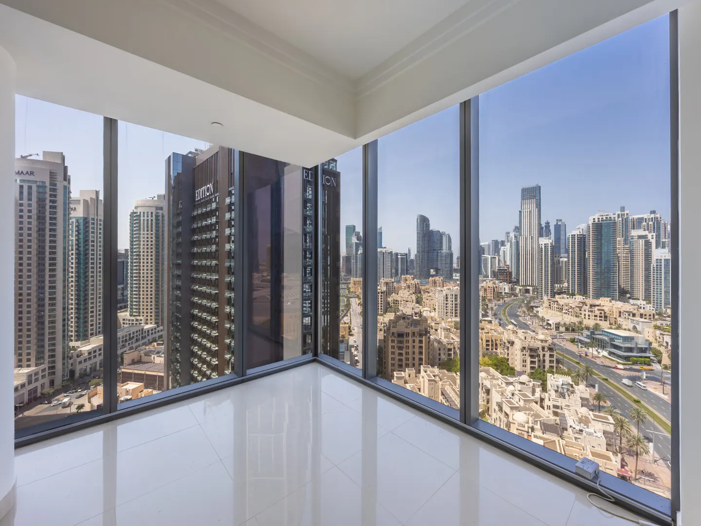 Corner view of a bright, empty room with white tile floors and floor-to-ceiling windows overlooking a cityscape with tall buildings.