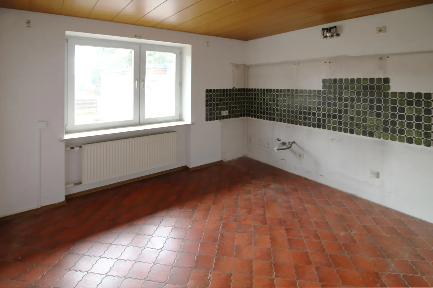 Empty kitchen with red tile floor, white walls, and wood ceiling. A window lets in light. Green and black tiles line the wall.