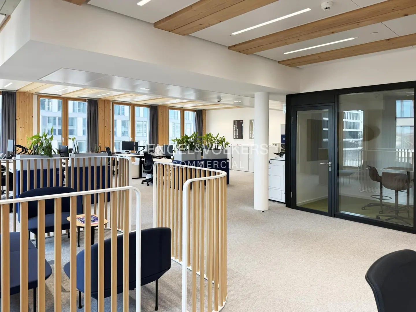Bright office space with wood accents, featuring blue chairs, desks, and a glass-walled conference room. Engel & Volkers logo visible.